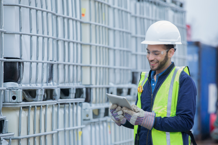Young adult Hispanic man is chemical engineer checking inventory shipment of chemical crates on oil and gas pipeline job site. Man is wearing safety gear and using a digital tablet.
